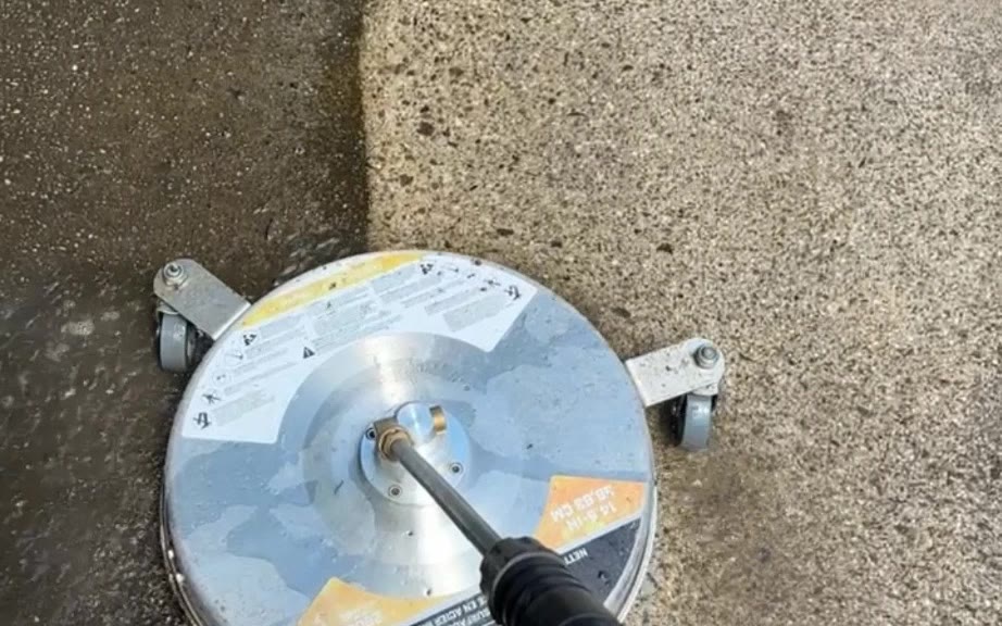 A rotary surface cleaner mid-pass on a driveway, with a hard line between the grey slab behind it and the clean concrete ahead